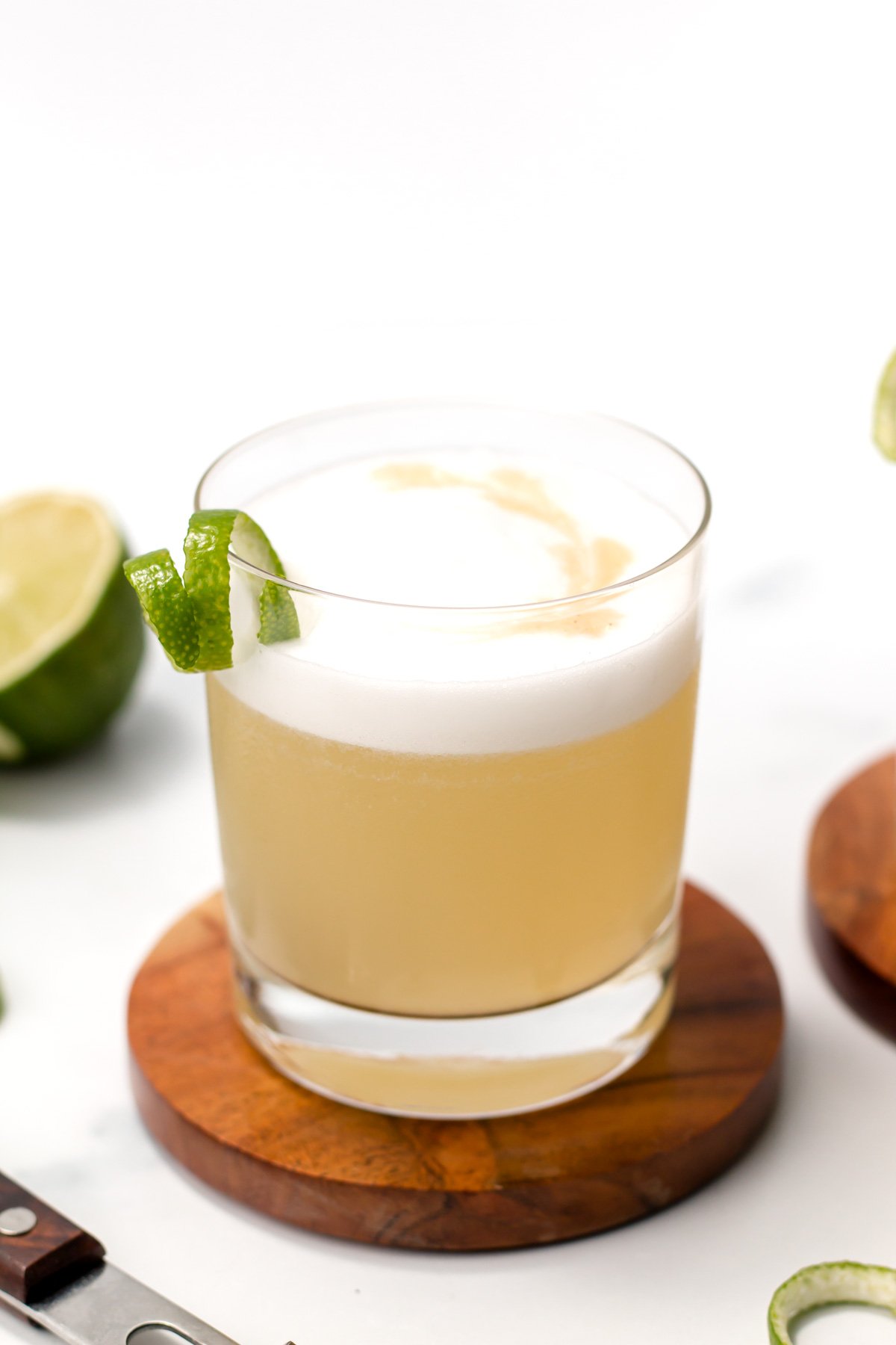 A glass of frothy cocktail garnished with a lime twist sits on a wooden coaster, with sliced limes and a knife nearby.