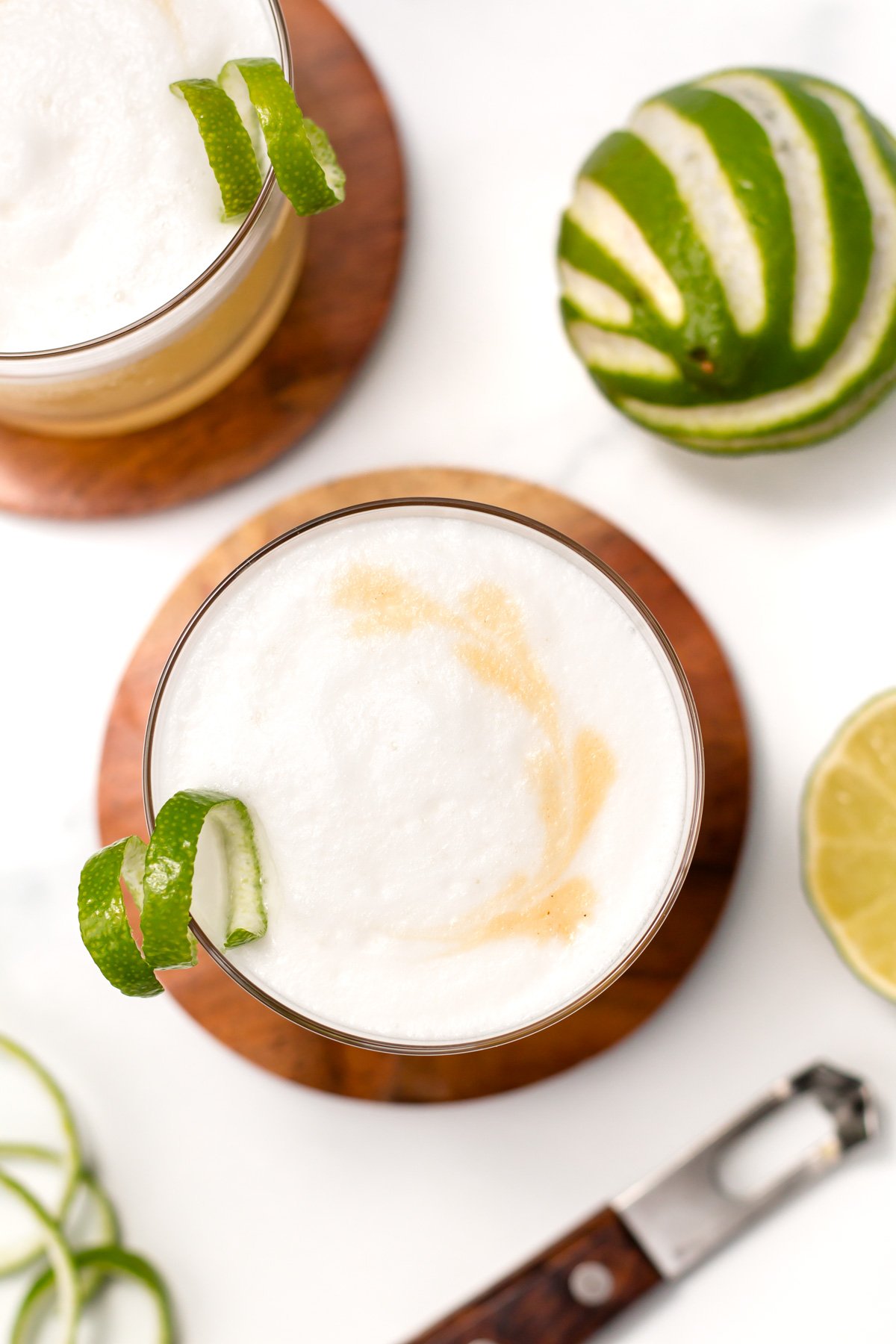 Overhead view of a cocktail with a foamy top, lime twist garnish, and a swirl of bitters, next to a zested lime, lime slice, and a zester on a white surface.