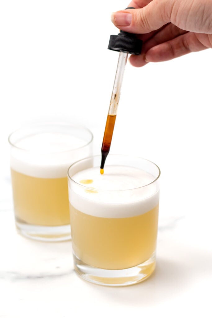 A hand uses a dropper to add liquid to a frothy yellow cocktail in a glass, with another similar drink in the background.