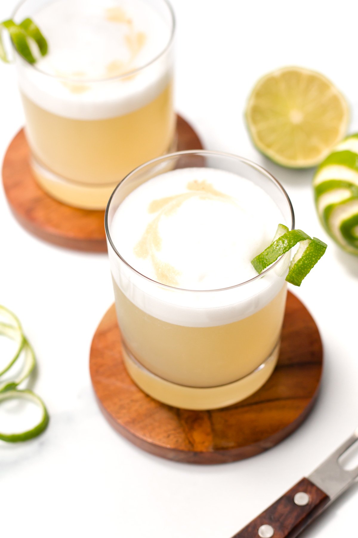 Two glasses of a light brown cocktail with frothy tops sit on wooden coasters, garnished with lime peel, next to a sliced lime and a knife.