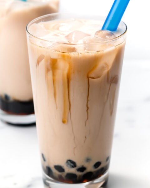 A glass of iced milk tea with black tapioca pearls at the bottom and a wide blue straw.
