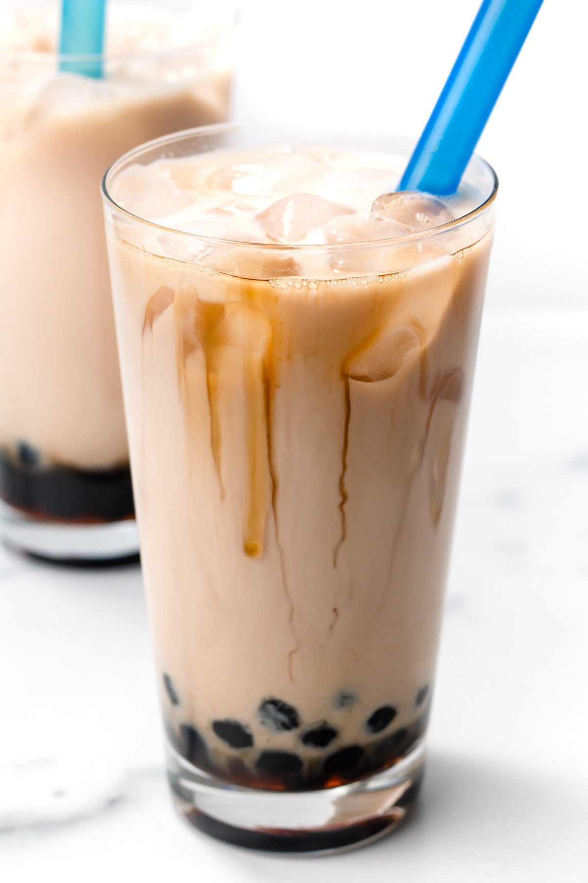 A glass of iced milk tea with black tapioca pearls at the bottom and a wide blue straw.