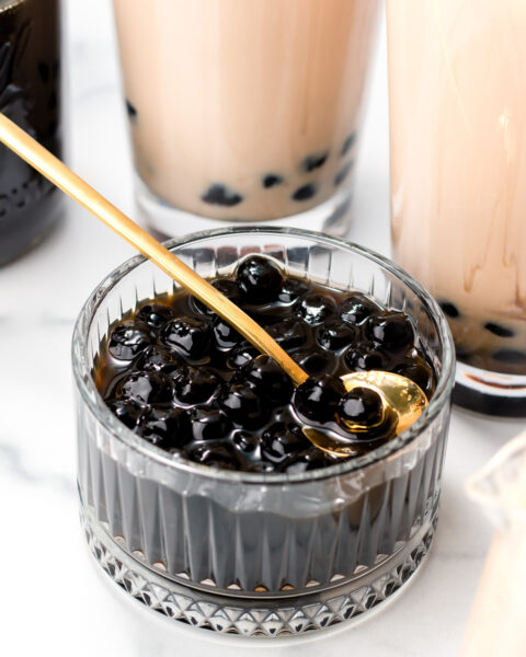 A small glass bowl filled with black tapioca pearls and syrup, with a golden spoon, surrounded by glasses of bubble tea on a white surface.