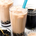 Iced bubble tea in tall glasses with blue straws, tapioca pearls at the bottom, and jars of black tea, extra pearls, and milk nearby.
