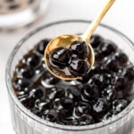 A gold spoon holds several black tapioca pearls above a glass bowl filled with more tapioca pearls, with a blurry bubble tea drink in the background.