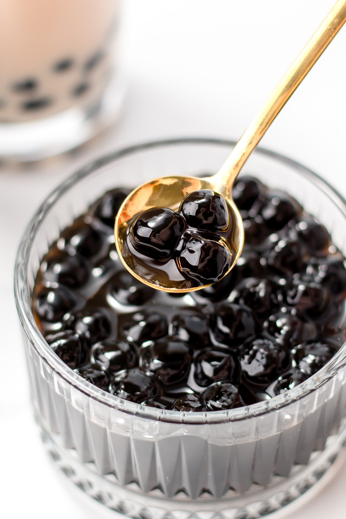 A gold spoon holds several black tapioca pearls above a glass bowl filled with more tapioca pearls, with a blurry bubble tea drink in the background.