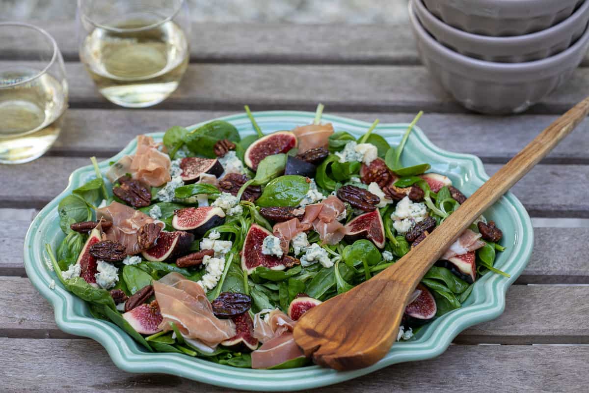A platter of spinach salad with figs, prosciutto, pecans, and crumbled cheese sits on a wooden table next to two glasses of white wine and stacked bowls.