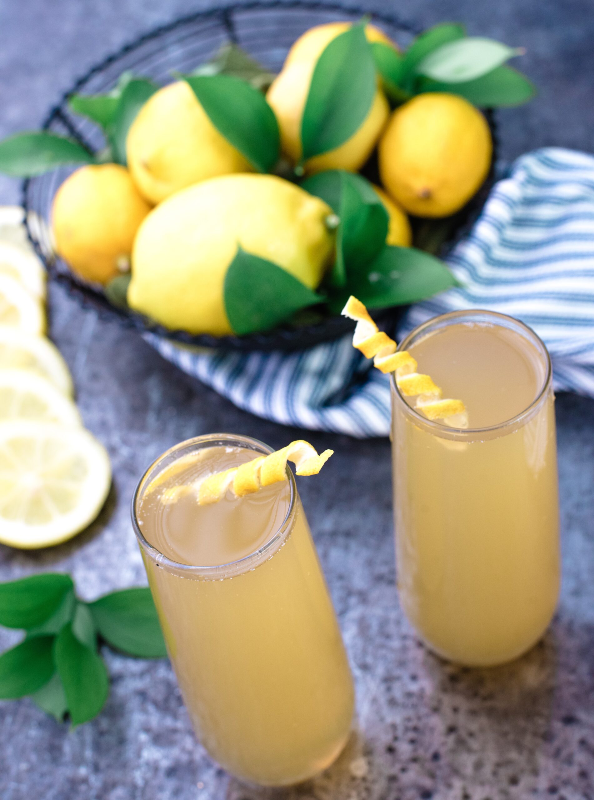 Two glasses of lemon drink with lemon peel garnish sit on a gray surface beside a basket of whole lemons, a striped cloth, and lemon slices.