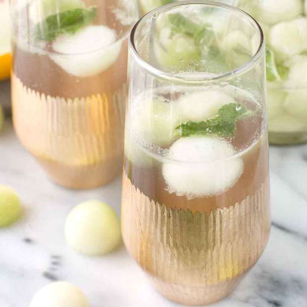 Two gold-rimmed glasses filled with a light beverage, ice cubes, mint leaves, and melon balls on a marble surface, with a jar of melon balls in the background.