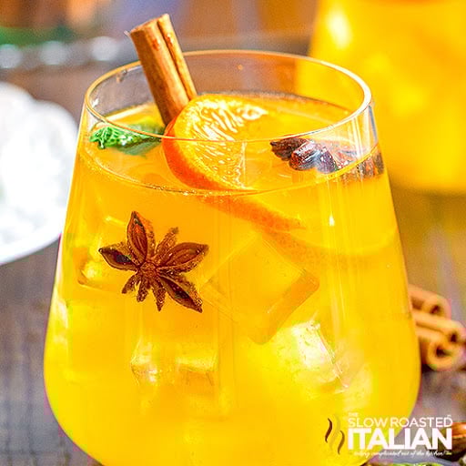 A glass of yellow-orange iced drink garnished with a cinnamon stick, orange slice, star anise, mint, and cloves.