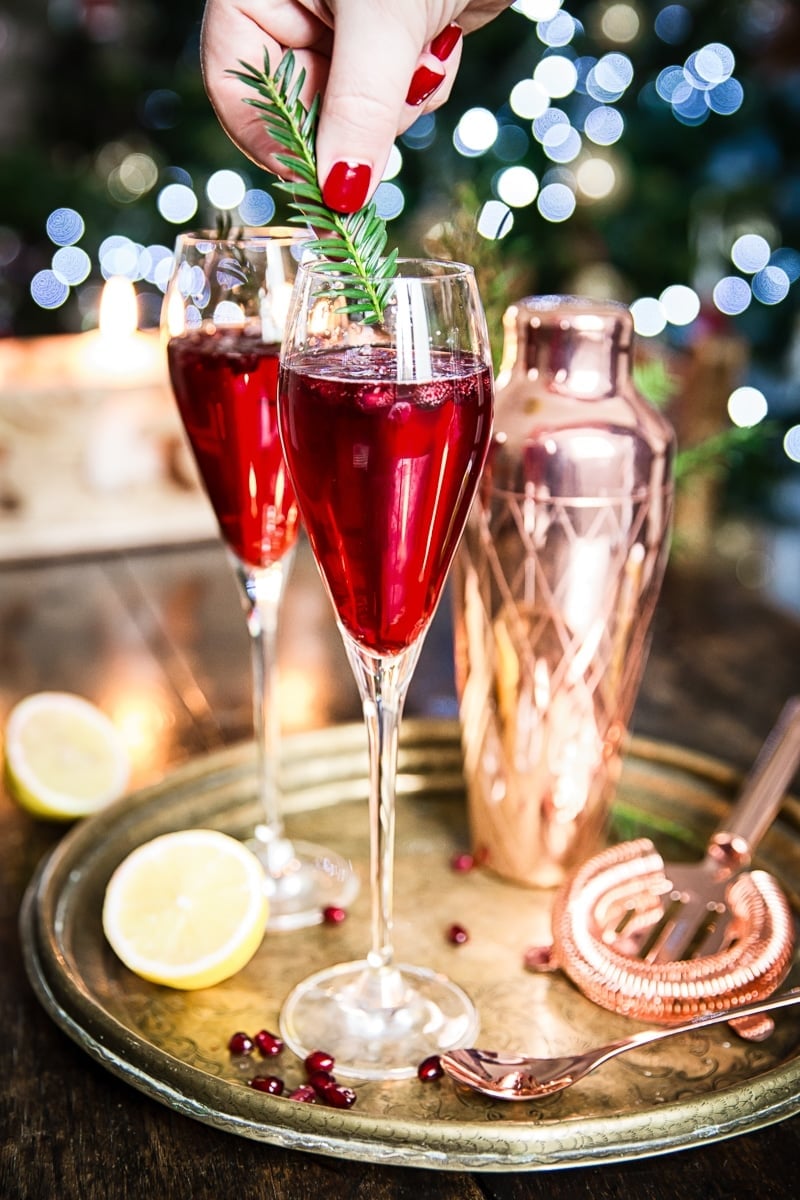A hand garnishes a red cocktail in a champagne flute with a rosemary sprig; another filled glass, a copper shaker, lemon, and pomegranate seeds are on the tray.