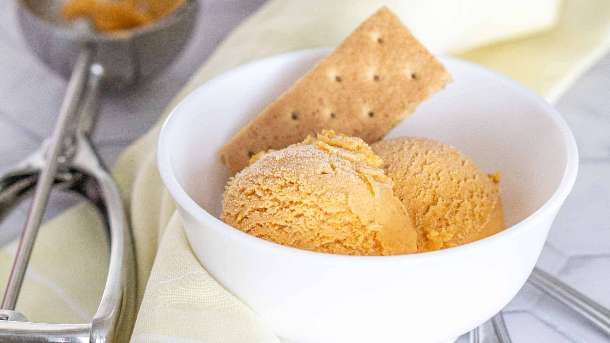 A white bowl with two scoops of orange ice cream and a rectangular biscuit, with an ice cream scoop and a cloth nearby.