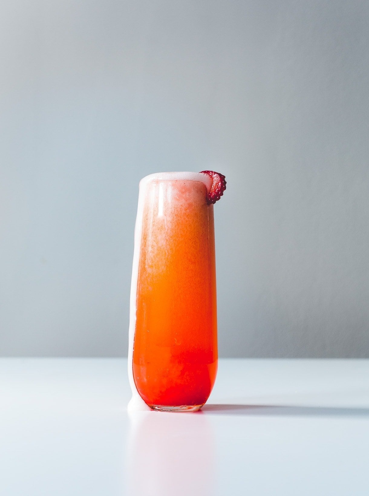 A tall glass of orange-red fizzy drink with foam on top, garnished with a slice of strawberry on the rim, placed on a white surface against a plain background.