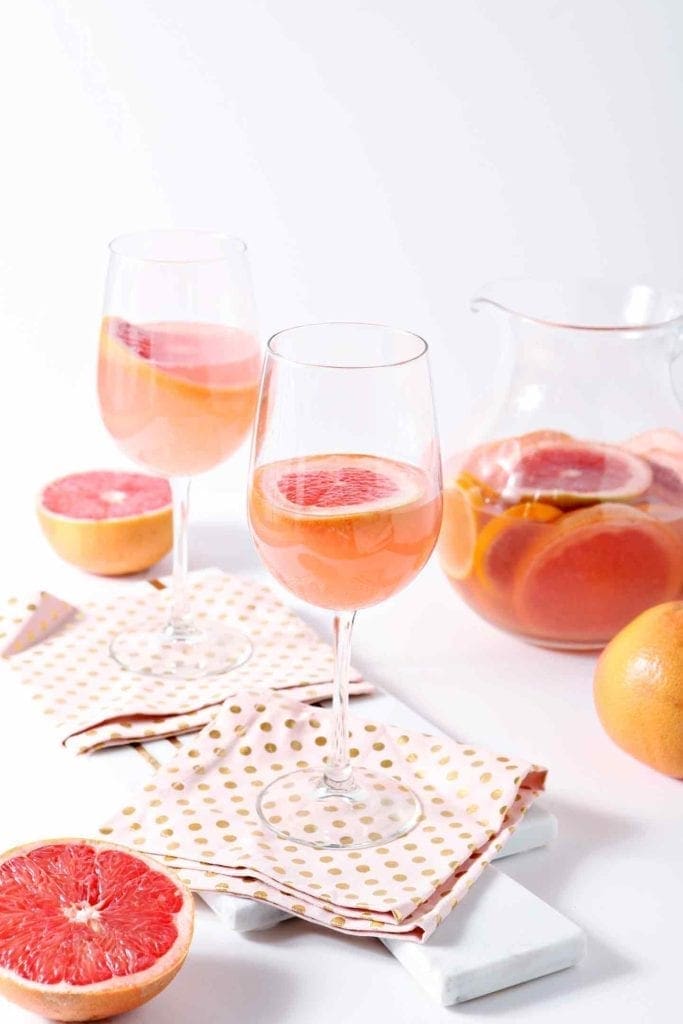 Two wine glasses and a pitcher contain a pink beverage with grapefruit slices, surrounded by halved grapefruits and polka dot napkins on a white surface.