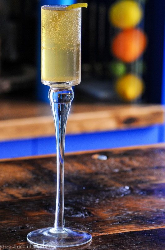 A tall, clear champagne flute with a light yellow sparkling drink and a lemon twist garnish, placed on a wooden surface.