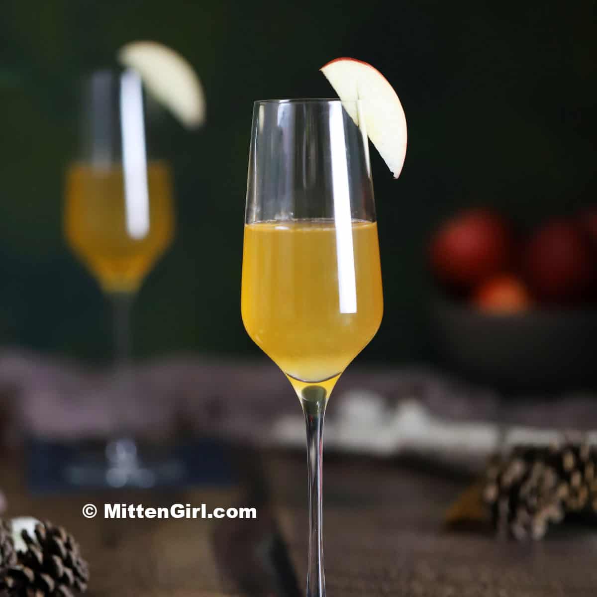 A champagne glass filled with a yellow-orange drink, garnished with an apple slice, sits on a table with pinecones; another glass and apples are blurred in the background.