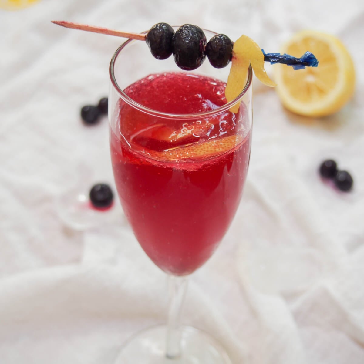 A glass of red cocktail garnished with a lemon twist and blueberries on a skewer, with lemon slices and blueberries in the background.