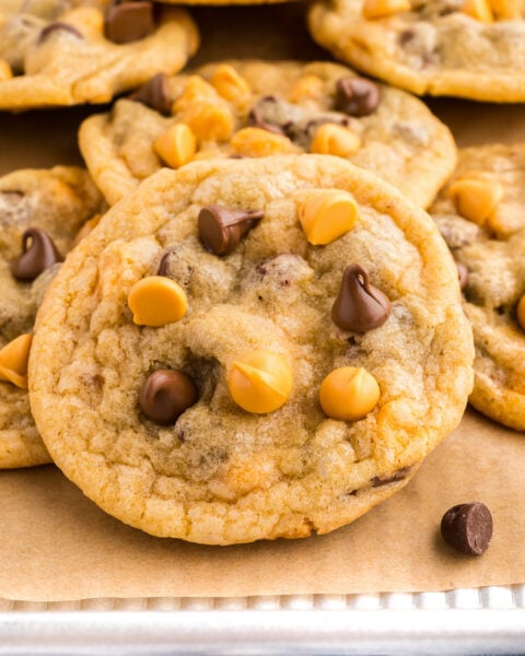 A close-up of several chocolate chip cookies with butterscotch and chocolate chips on a parchment-lined baking tray.