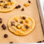 A baking sheet lined with parchment paper holds several chocolate chip cookies topped with both dark and butterscotch chips, with extra chips scattered around.