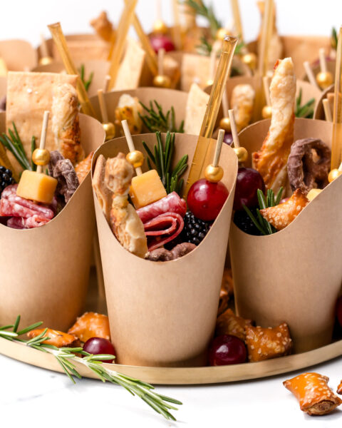 Individual brown paper cups filled with assorted charcuterie items including cheese cubes, salami, crackers, grapes, blackberries, and rosemary sprigs, arranged on a tray.