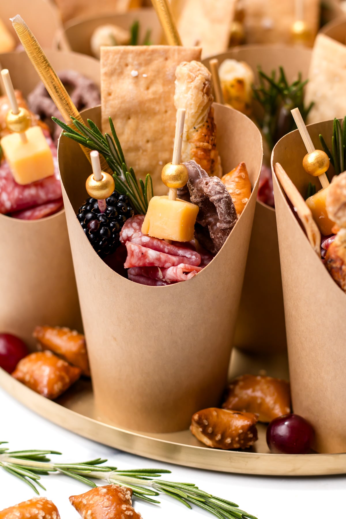 Brown paper cups filled with skewered cheese, cured meat, crackers, blackberries, and pretzels, garnished with rosemary, arranged on a tray.