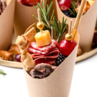A paper cone filled with assorted charcuterie items including cheese, salami, grapes, blackberries, a breadstick, pretzel, and a rosemary sprig.