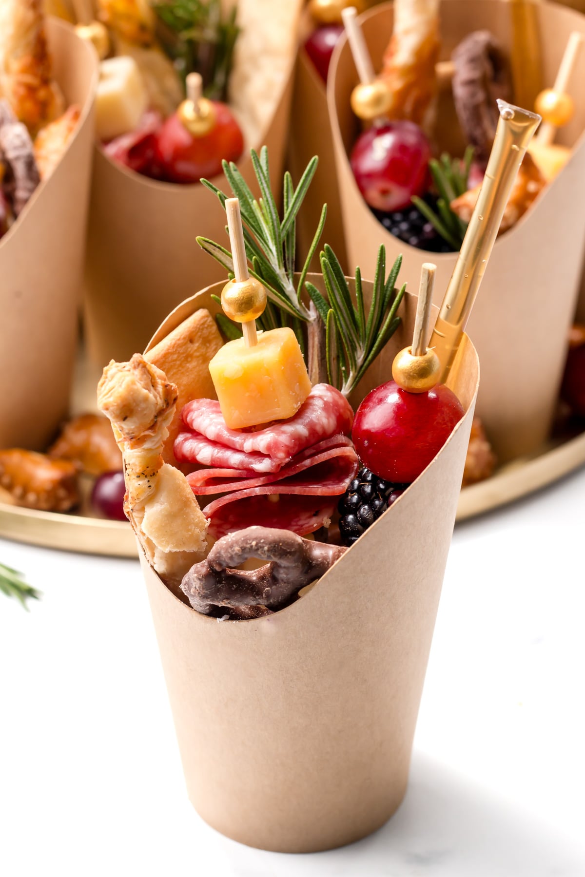 A paper cone filled with assorted charcuterie items including cheese, salami, grapes, blackberries, a breadstick, pretzel, and a rosemary sprig.