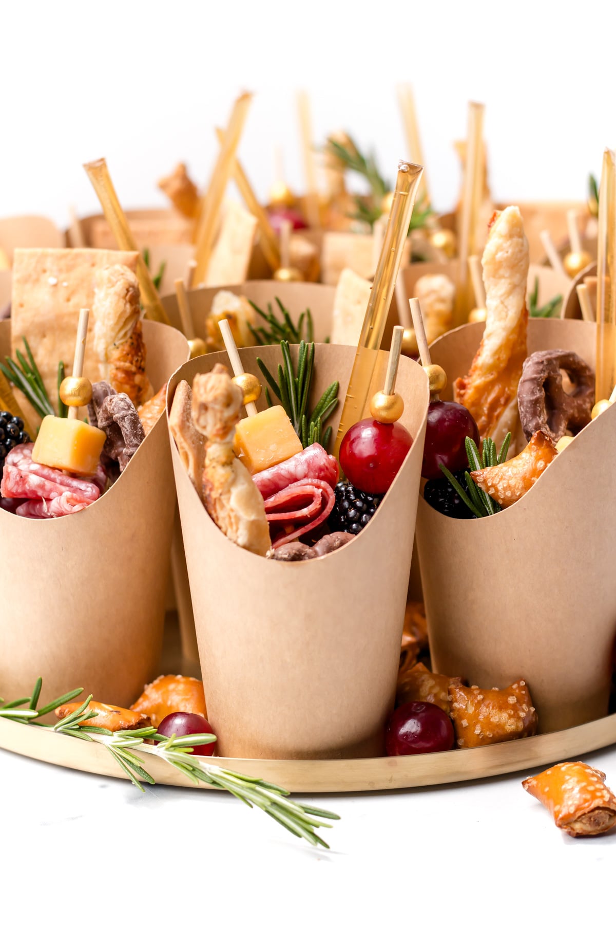 Brown paper cups filled with assorted snacks, including cheese, salami, grapes, crackers, rosemary sprigs, and breadsticks, arranged on a round tray.