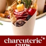 A paper cup filled with assorted charcuterie items including cured meats, cheese, grapes, berries, nuts, and a breadstick, garnished with rosemary.