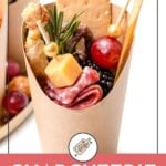 A paper cup filled with salami, cheese, crackers, grapes, olives, a breadstick, and berries, with a toothpick and garnish. Text reads: "Charcuterie Cups.