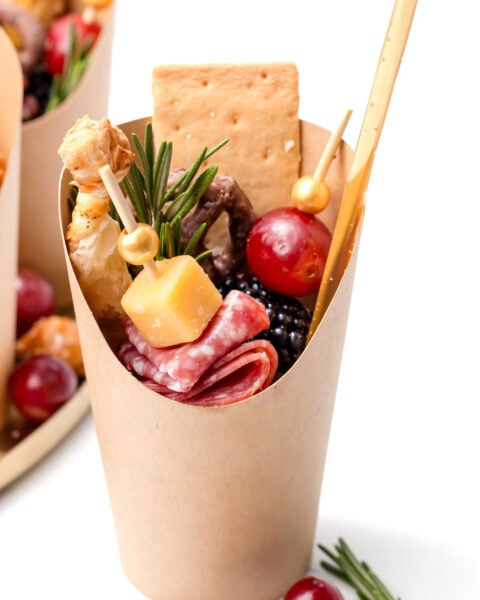 A paper cup filled with salami slices, cheese cubes, a cracker, grapes, blackberries, a breadstick, and a sprig of rosemary, arranged for individual serving.