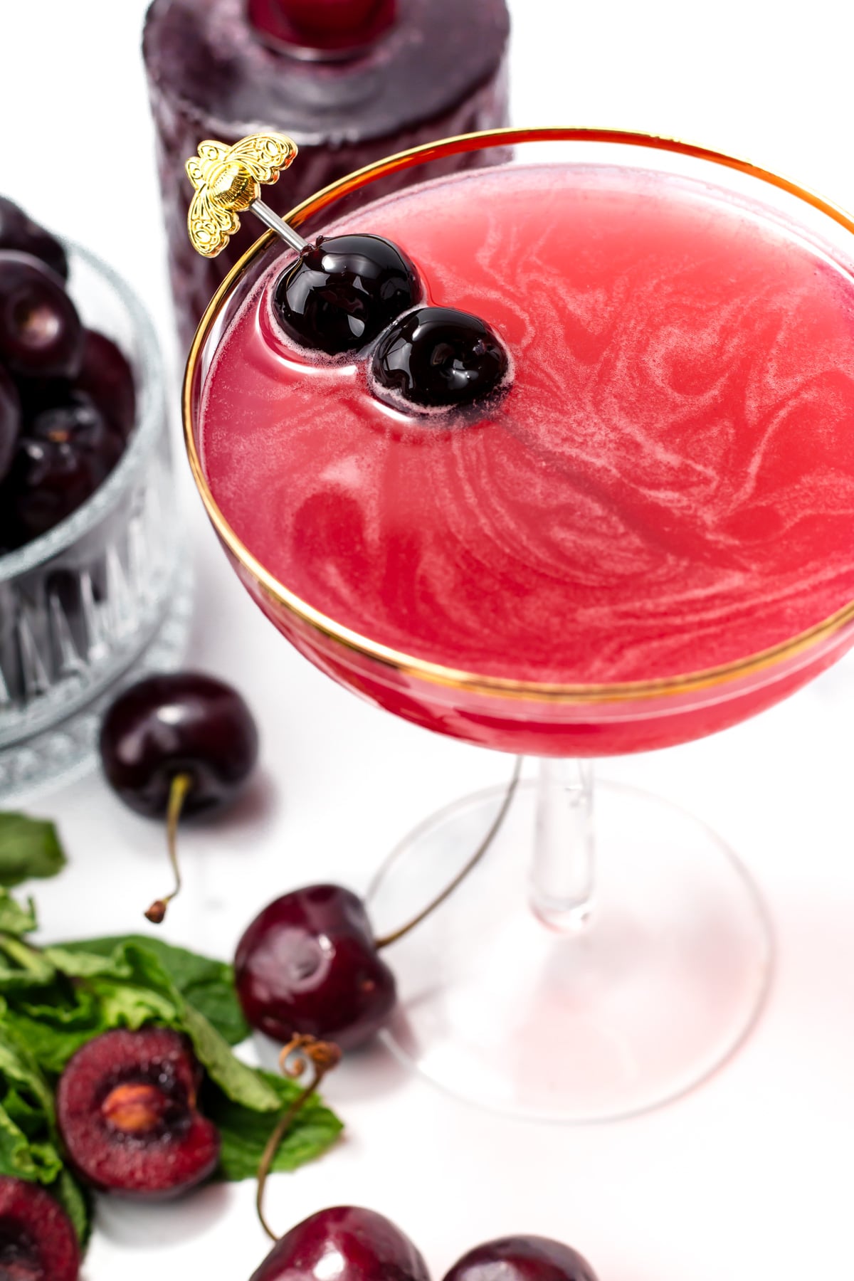 A pink cocktail garnished with two cherries on a gold pick, surrounded by fresh cherries and mint leaves on a white surface.