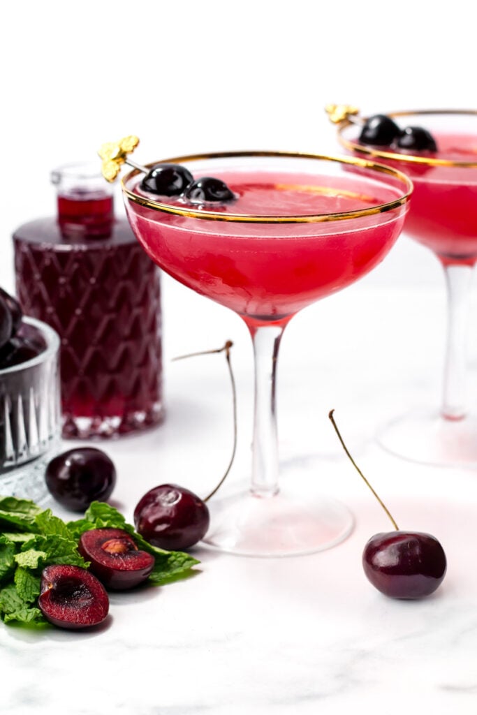 A coupe glass filled with a bright red cherry cocktail, garnished with whole cherries; nearby are fresh cherries, mint leaves, and a glass bottle of red liquid on a white surface.