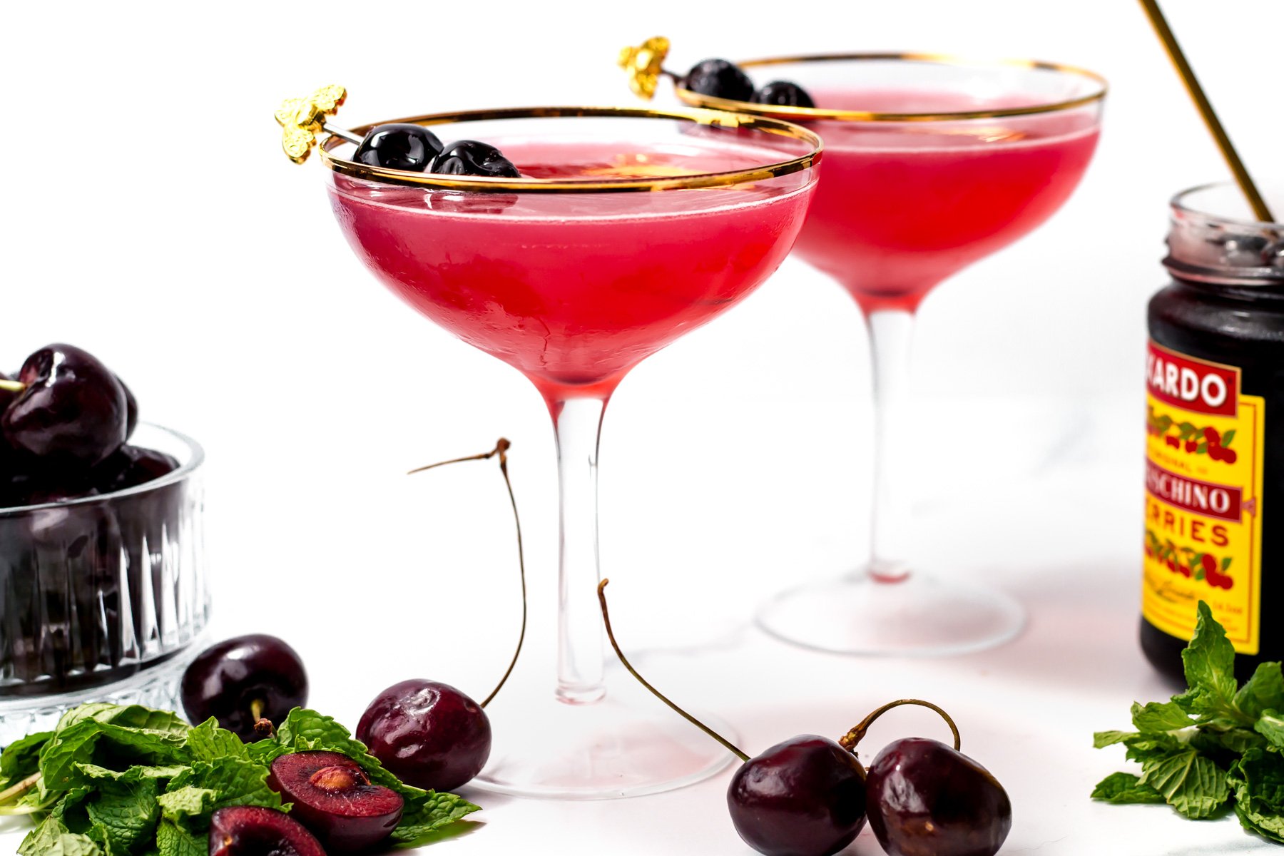 Two coupe glasses filled with a red cocktail garnished with cherries, surrounded by fresh cherries, mint leaves, and a jar of maraschino cherries on a white surface.