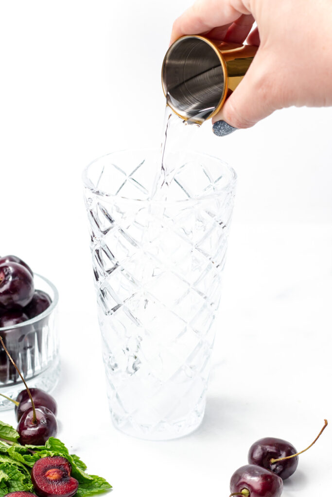 A hand pours liquid from a metal jigger into a patterned glass, with cherries and mint leaves nearby on a white surface.