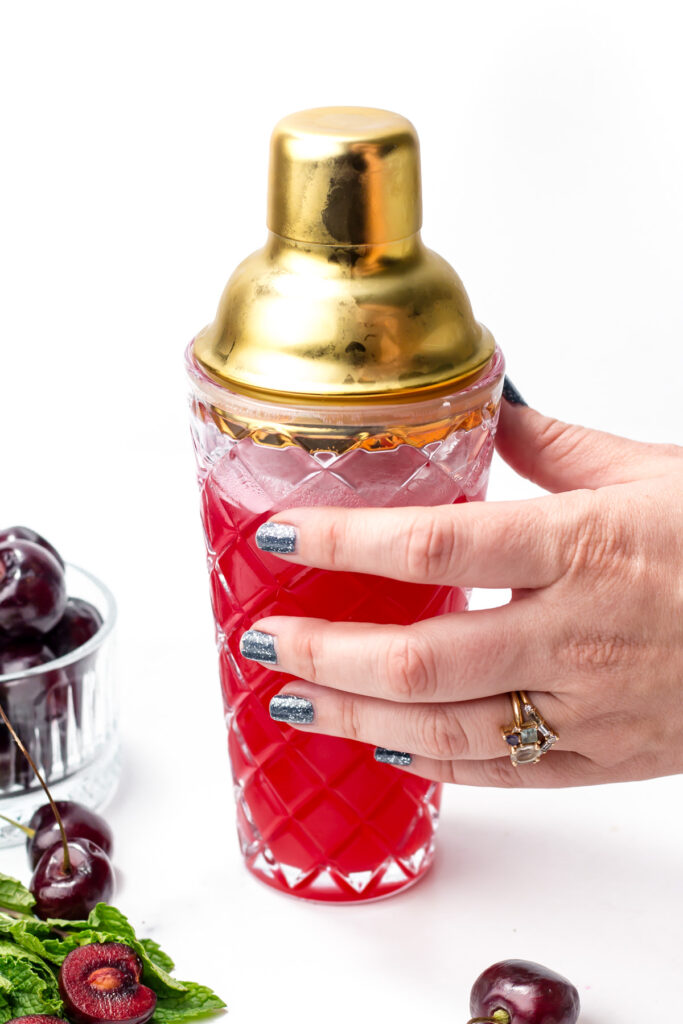 A hand with painted nails holds a crystal cocktail shaker filled with a red drink, surrounded by fresh cherries and mint on a white surface.
