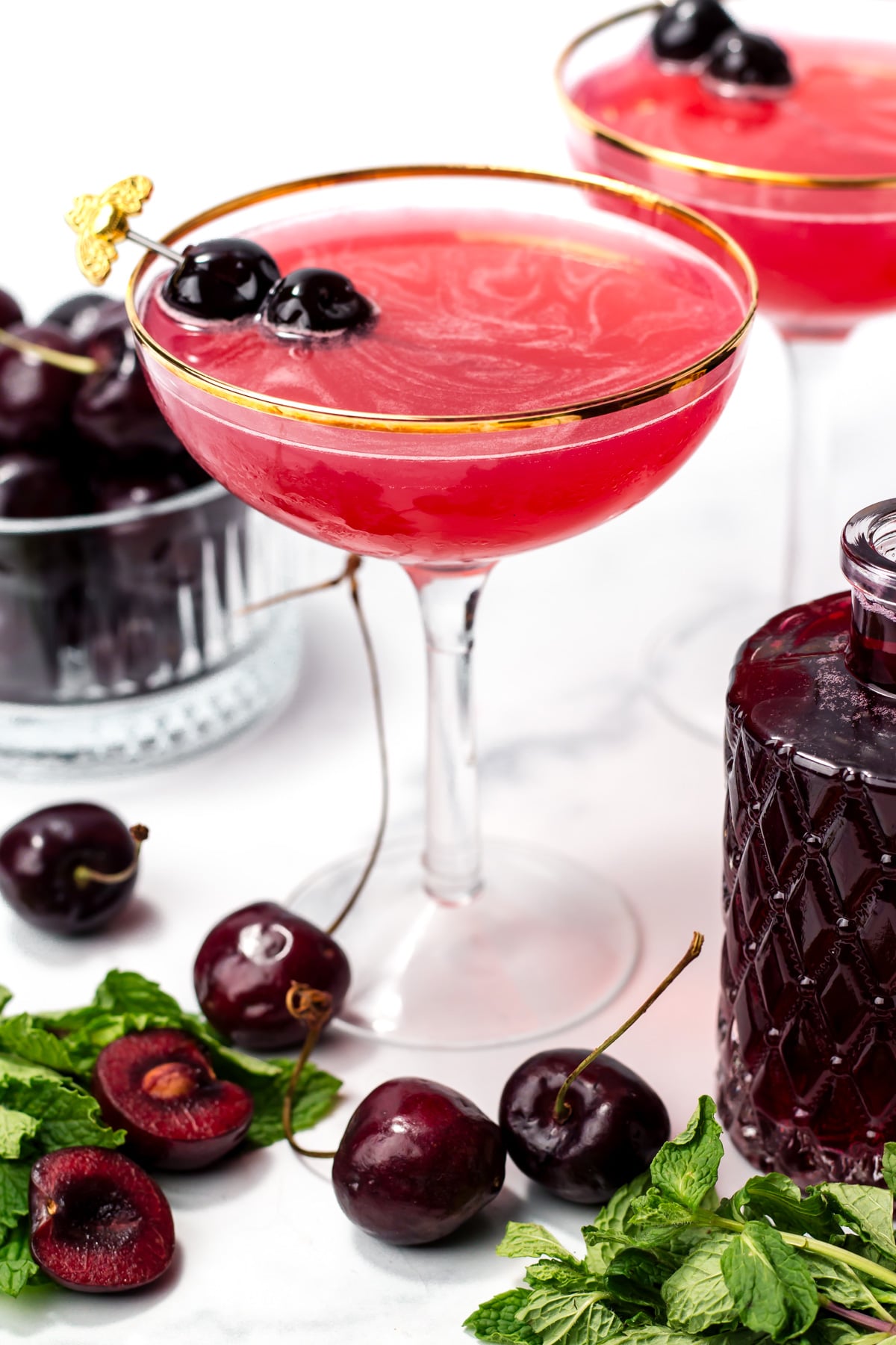 A coupe glass filled with a bright red cocktail garnished with two cherries, surrounded by fresh cherries, mint leaves, and a glass bowl of whole cherries.