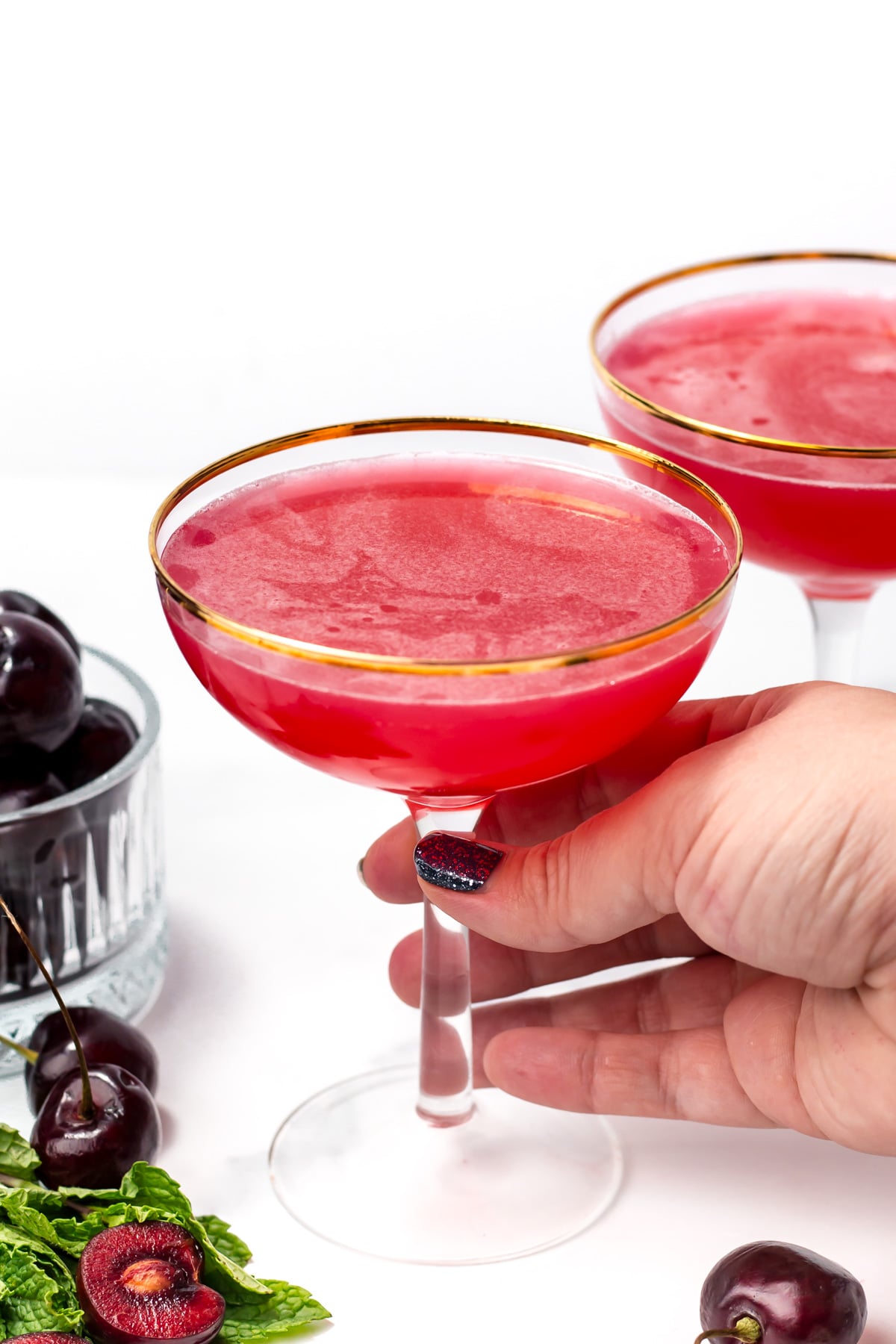A hand holding a coupe glass filled with a bright red cocktail; cherries and mint leaves are arranged nearby, with a second glass in the background.