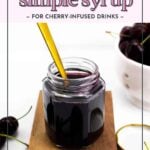 A jar of dark cherry simple syrup with a golden spoon, placed on a wooden board, surrounded by fresh cherries.