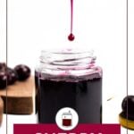 An open jar filled with dark cherry syrup sits on a white surface, with a drop of syrup falling into it. Cherries and a wooden board are visible in the background.
