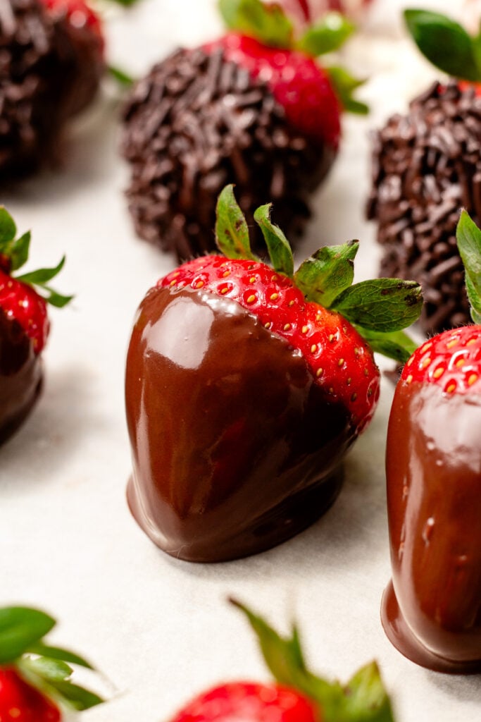 Close-up of fresh strawberries partially dipped in chocolate, arranged on a white surface. Some strawberries in the background are coated with chocolate sprinkles.