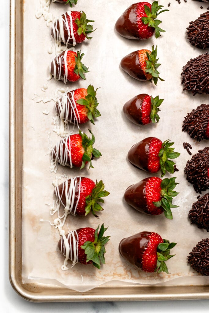 Rows of strawberries dipped in chocolate, some drizzled with white chocolate, arranged on parchment paper next to chocolate-covered treats on a baking sheet.