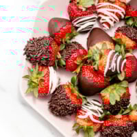 A plate of strawberries dipped in chocolate, some with chocolate sprinkles and others decorated with white chocolate drizzle.