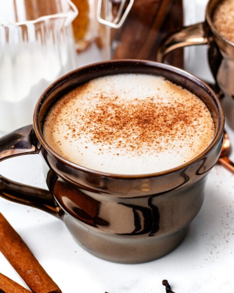A brown ceramic mug filled with a frothy, spiced latte topped with ground cinnamon, surrounded by cinnamon sticks and vanilla beans on a white surface.