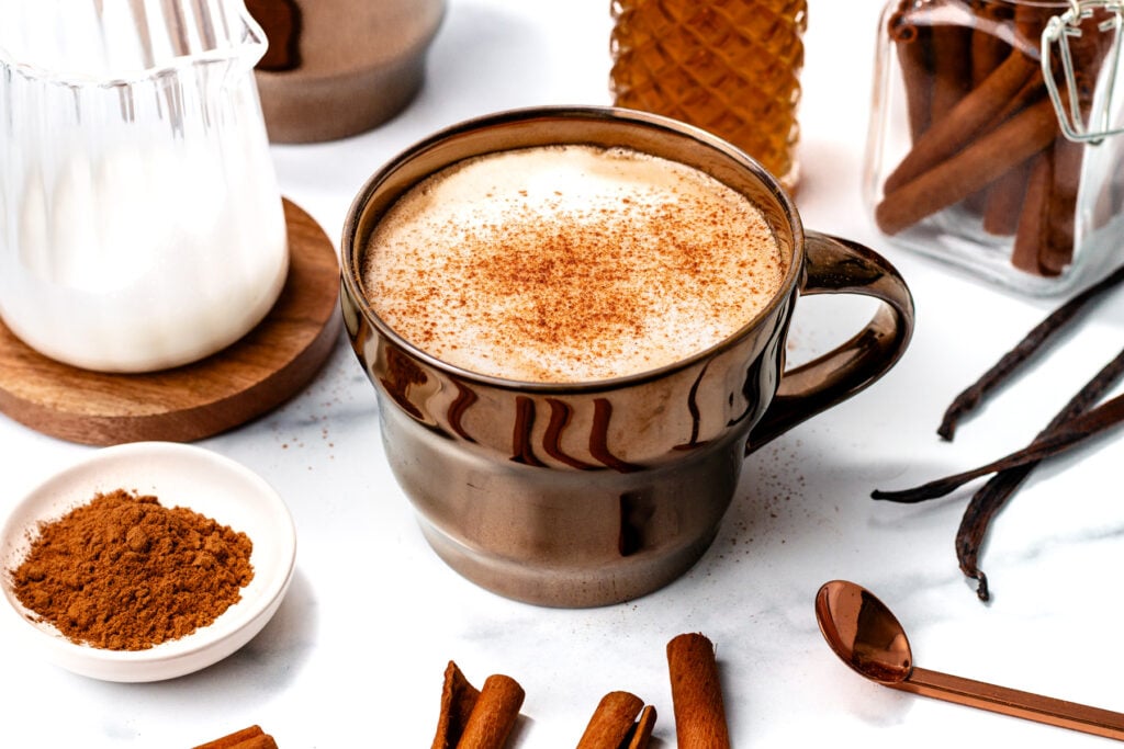 A brown ceramic mug filled with a foamy spiced latte, topped with cinnamon, surrounded by cinnamon sticks, a spice jar, milk, and a spoon on a white surface.