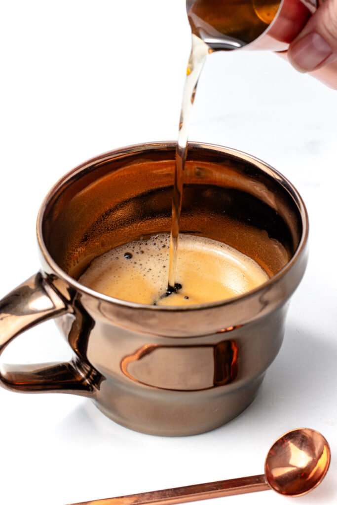 A hand pours liquid from a small pitcher into a cup of coffee, with a spoon resting nearby on a white surface.