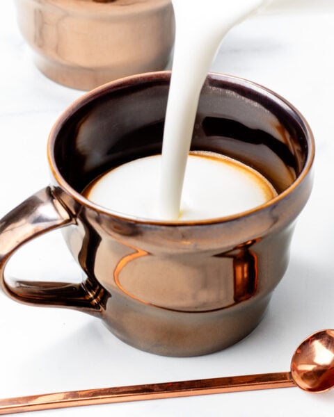 Milk is being poured into a metallic brown mug, with another similar mug and a matching spoon nearby on a white surface.