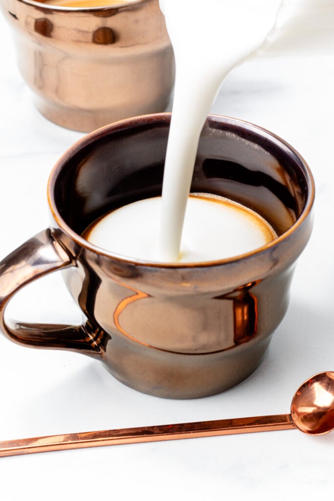 Milk is being poured into a metallic brown mug, with another similar mug and a matching spoon nearby on a white surface.