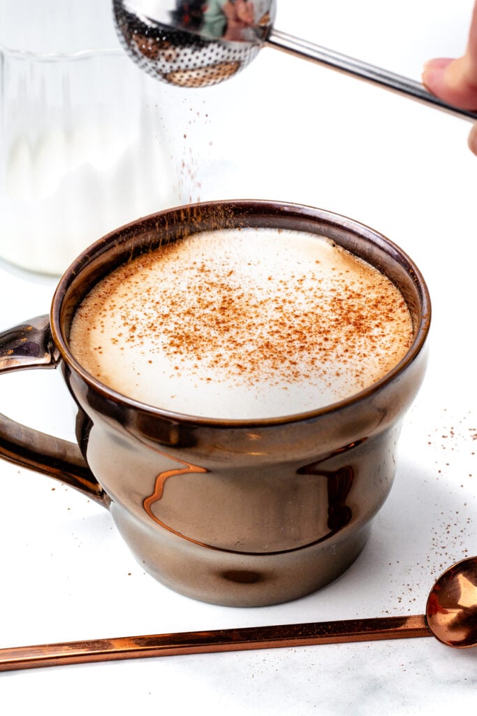 A frothy coffee drink in a metallic mug is topped with cocoa powder, with a sieve and a spoon nearby on a white surface.