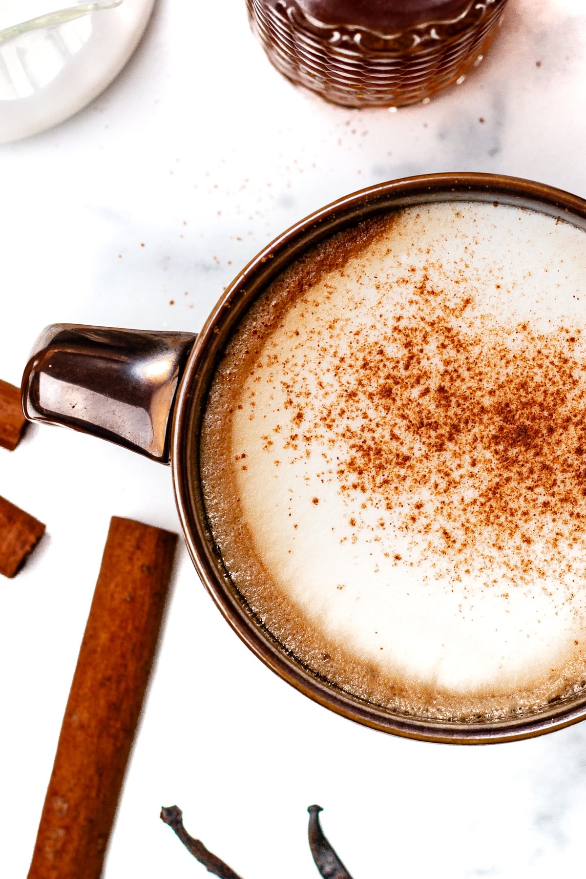 A brown ceramic mug filled with frothy coffee topped with cinnamon, surrounded by cinnamon sticks and a spice on a white surface.